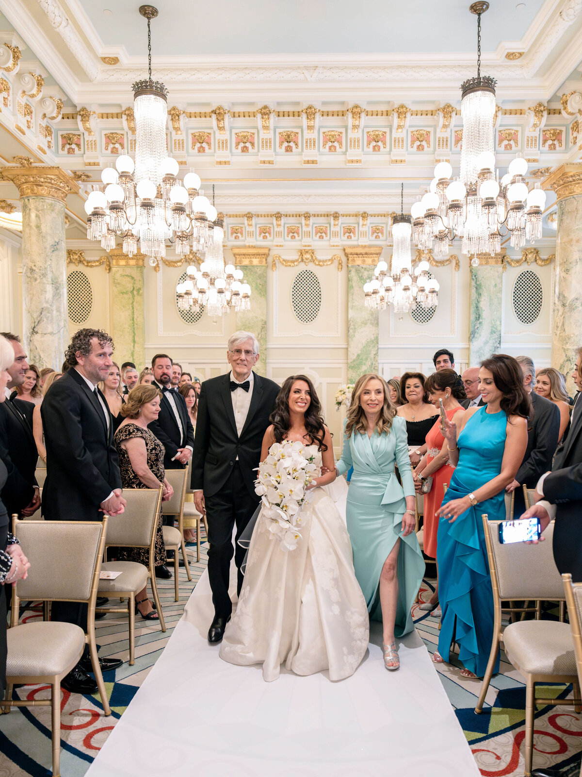 A bride in a white gown walks down the aisle with two companions in an ornate room with chandeliers. Guests are seated on either side, watching the procession. The setting is elegant, with intricate decor and a blue and white carpet.