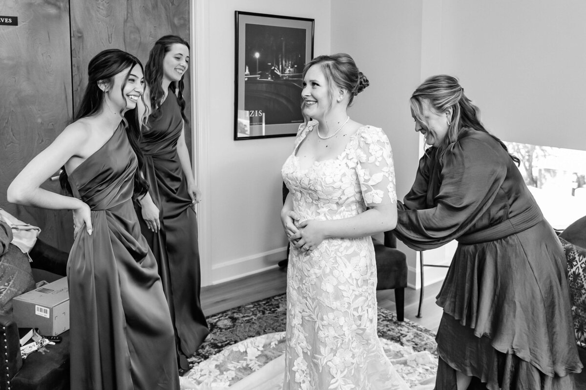 A bride in a lace gown smiles as three women in dark dresses help her get ready. They stand together in a bright room, laughing and enjoying the wedding preparations.