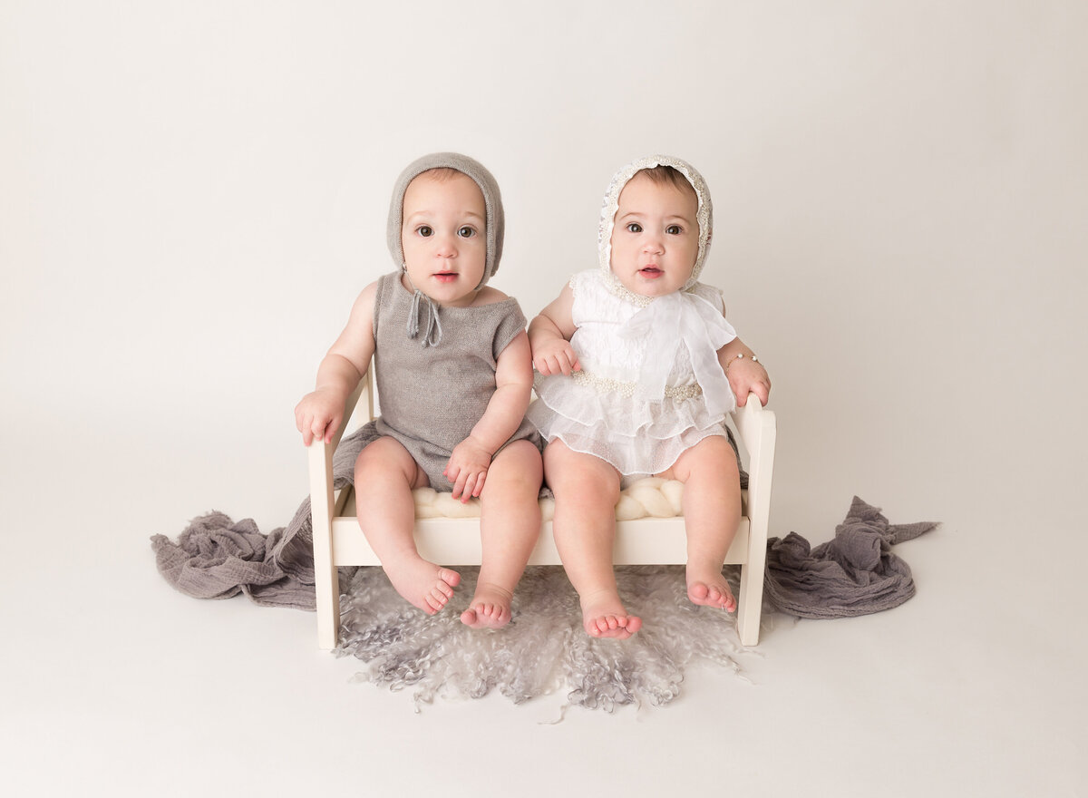 Twin baby girl and boy are sitting on a tiny bed for a 6-month milestone photoshoot. Babies are looking at the camera.