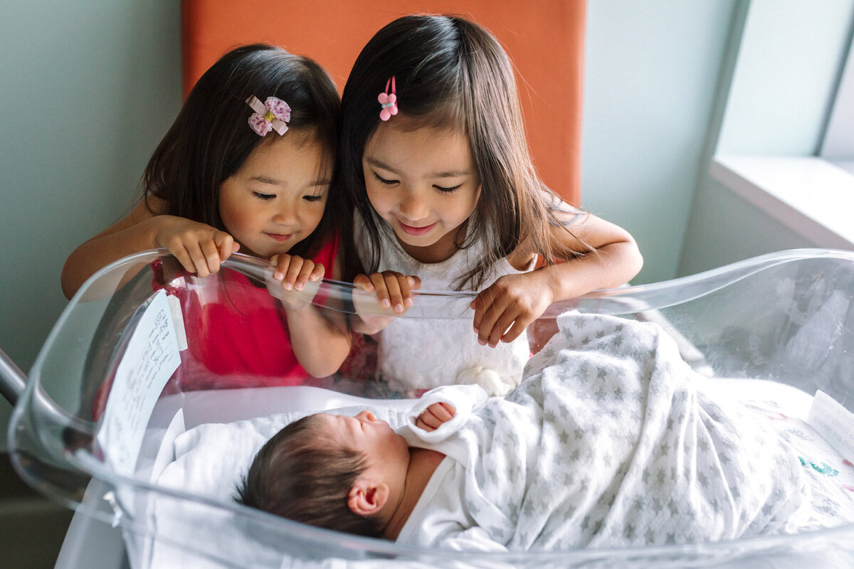 sisters standing and looking into hospital bassinet and newborn brother