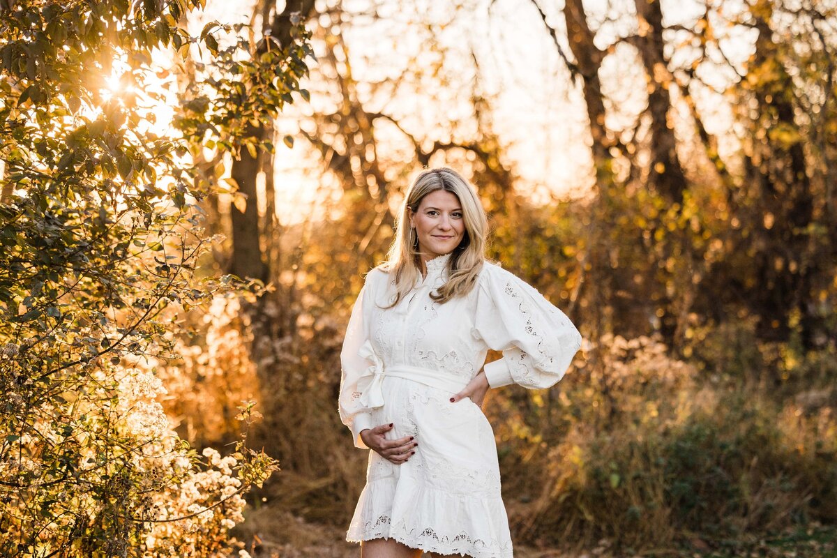 Woman in a white dress standing in a sunlit autumnal forest for her maternity photography session.