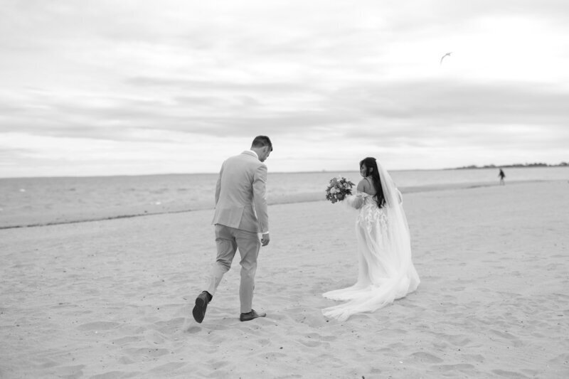 A couple walks side by side on a sandy beach, captured beautifully by a New England elopement photographer. The groom wears a light suit, and the bride, in a flowing dress and veil, holds her bouquet. An overcast sky looms as the ocean whispers in the background, with a bird gliding overhead.