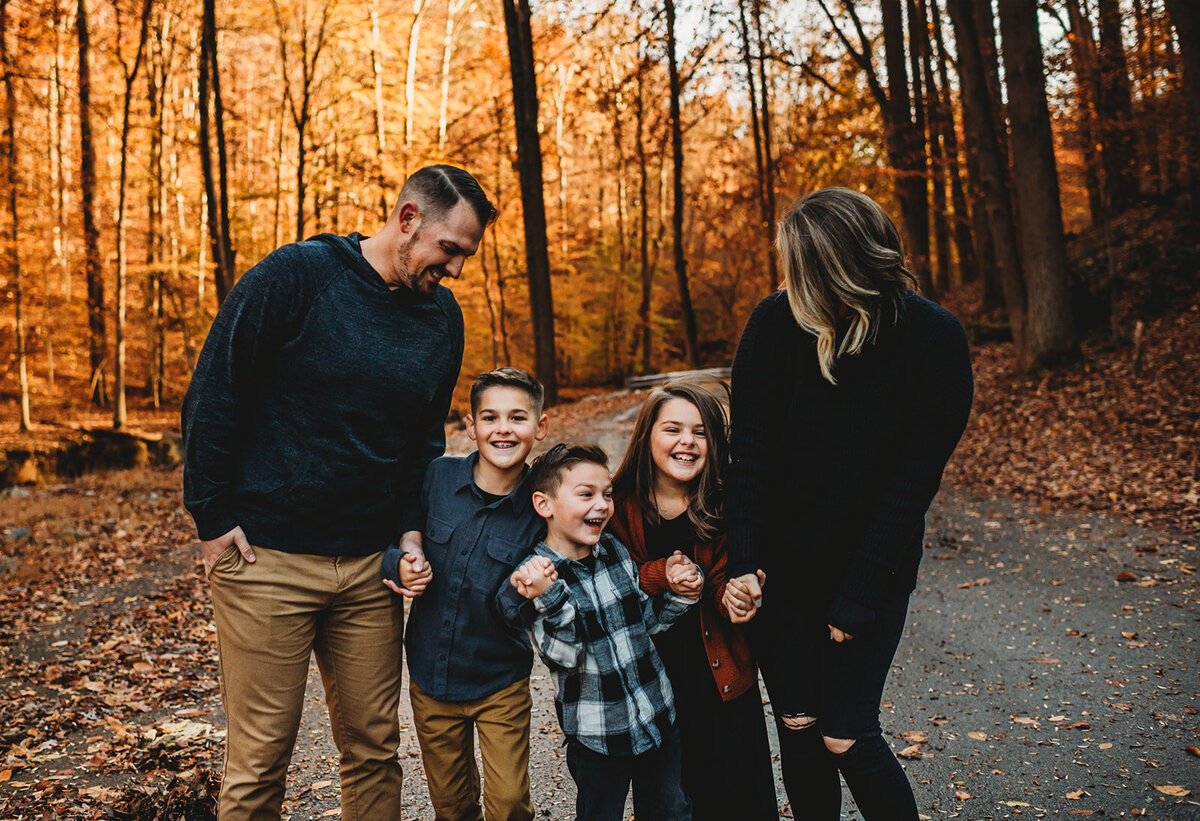 Baltimore photographers captures fall famil pictures with parents holdin ghands together with their three little children as they walk on a trail together with the woods in the distance with autumn colors