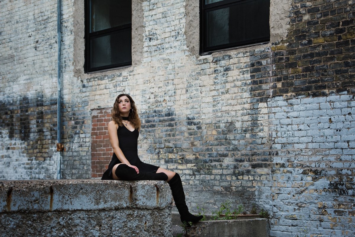 high school senior girl with knee high fashion boots sitting on concrete in front of urban brick wall
