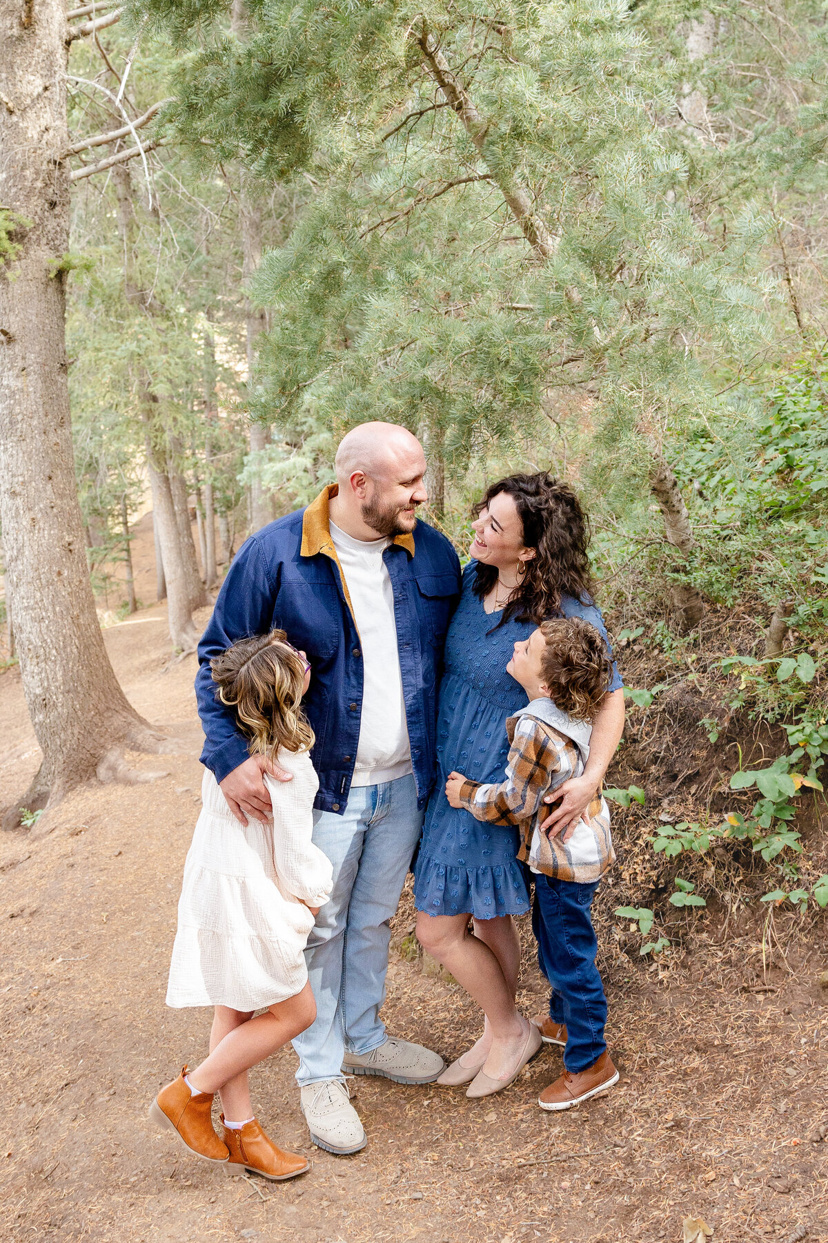 A candid moment of a family of four standing closely together in a wooded area, sharing smiles and laughter while surrounded by tall pine trees.