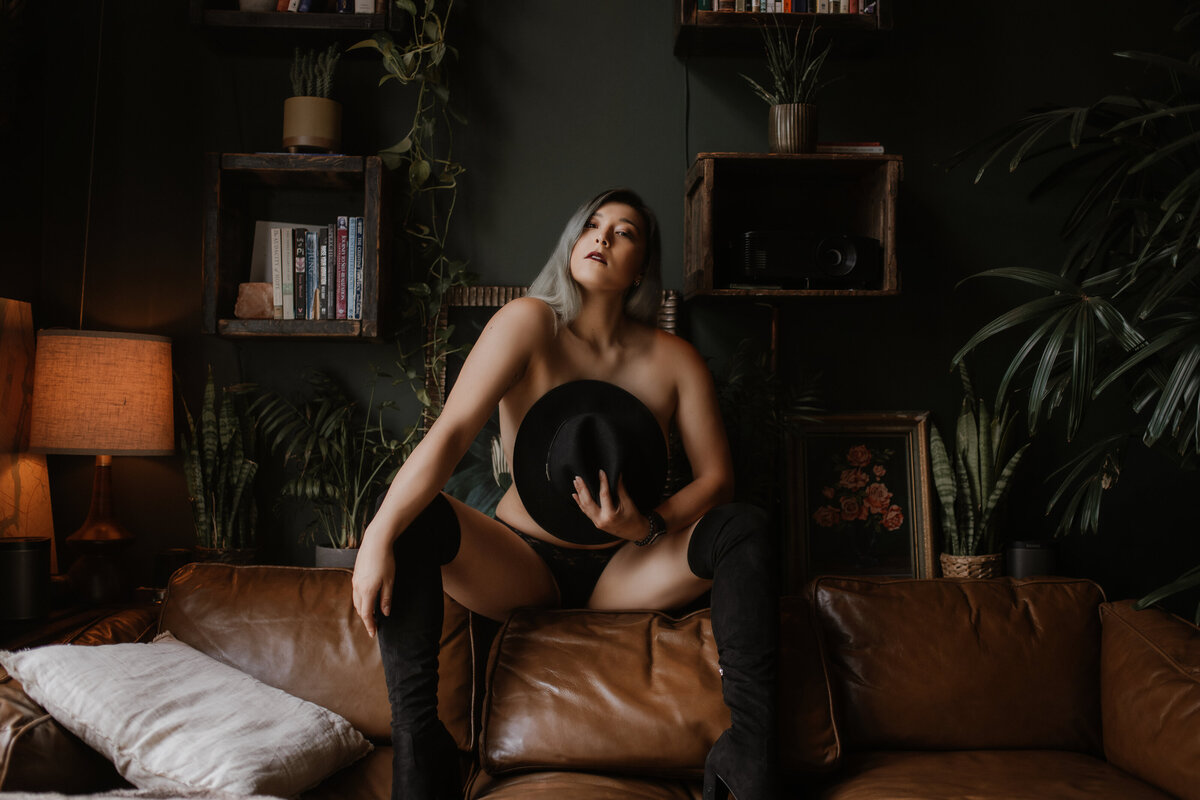 A woman wearing nothing but black boots covers her chest with a hat while posing on a couch during a boudoir session in Vancouver, BC.