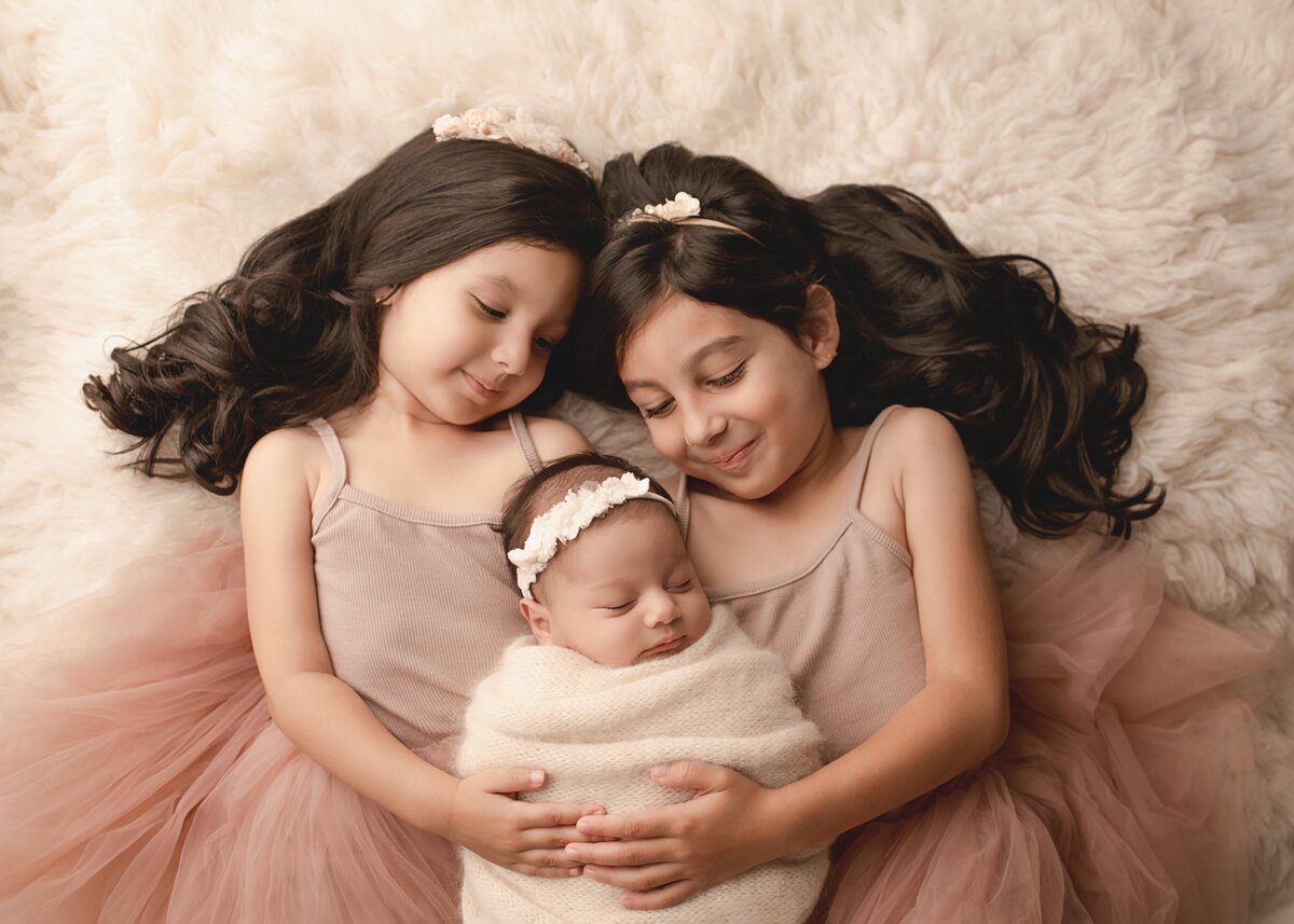 Aerial image. Newborn baby girl is captured with her two big sisters for her Lake Elsinore newborn photoshoot. The baby is laying between the two big sisters, who each have their hands gently resting atop of baby's belly. The baby is sleeping and wearing a white headband and cream knit swaddle. The big sisters have their heads touching and angled toward their new baby sister. Captured by best Lake Elsinore newborn photographer Bonny Lynn Photography