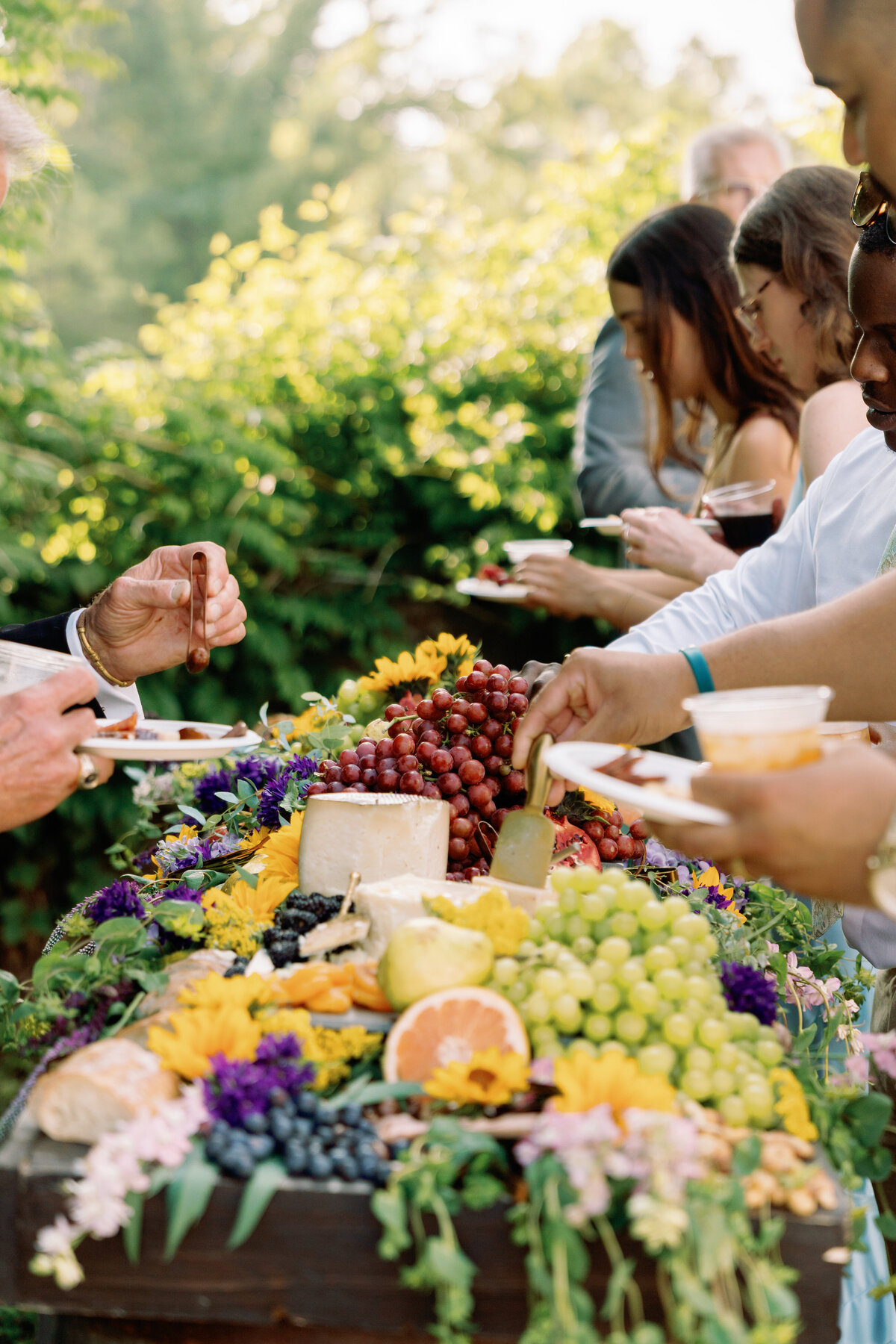 Guests during a wedding cocktail hour gathering plates of food at a charcuterie board. 