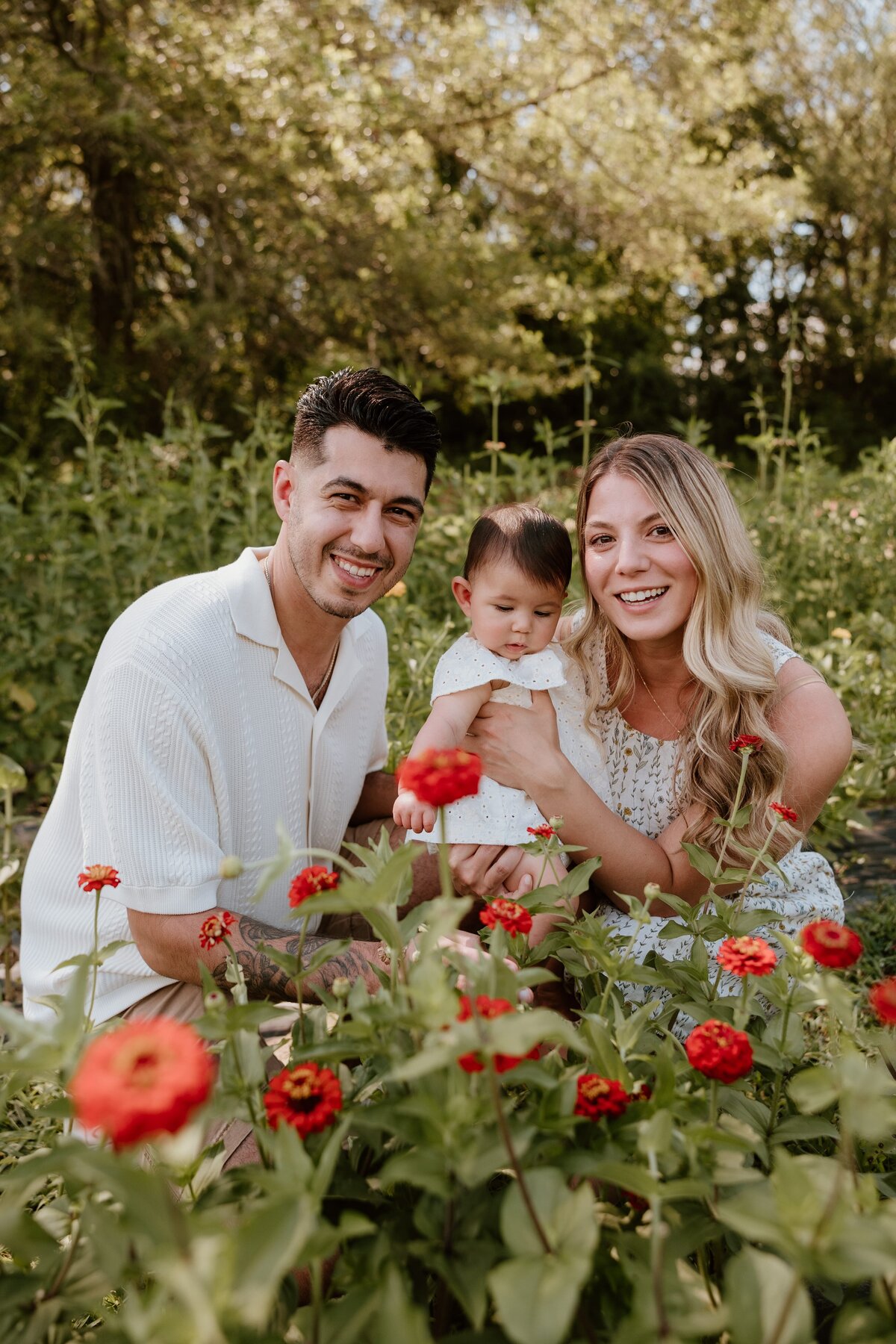 A happy family of three sits among vibrant red flowers at Brittany Hollow Farm in Rhinebeck. The father, wearing a white shirt, smiles brightly at the camera.