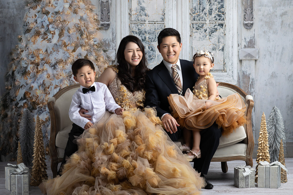 A Dallas family photographer captures a holiday portrait of a family of four. The mother and daughter wear matching golden dresses, while the father and son complement with formal attire. The background features a festive tree and holiday decor, creating a warm, loving atmosphere.