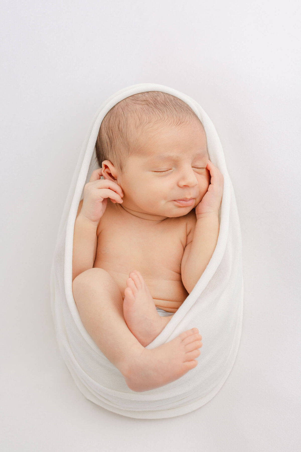 Newborn baby wrapped in a white swaddle up around his head. Hands are up by his face and his feet are hanging out a bit. He is sleeping during his minimalist all-white newborn photo session at Ashlie Behm Photography studios.