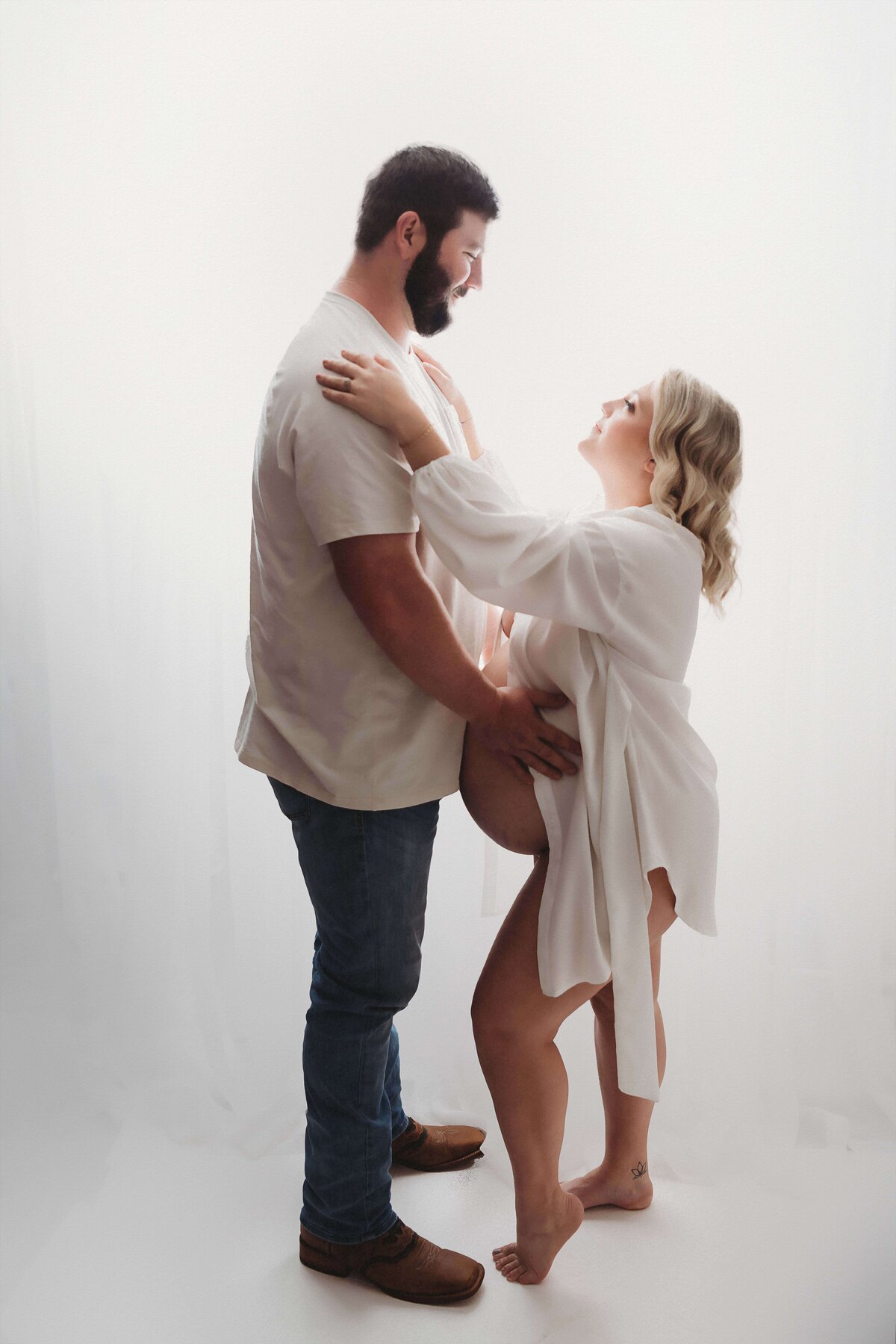 A young couple poses for a backlit maternity session with Mobile, AL Maternity Photographer. The dad is wearing his boots, blue jeans, and a white shirt as he embraces his wife's growing baby bump. She is wearing only a white button up shirt, which has been left unbuttoned to showcase her gorgeous belly and feminine figure. The two of them make eye contact, to create a romantic and timeless maternity portrait.