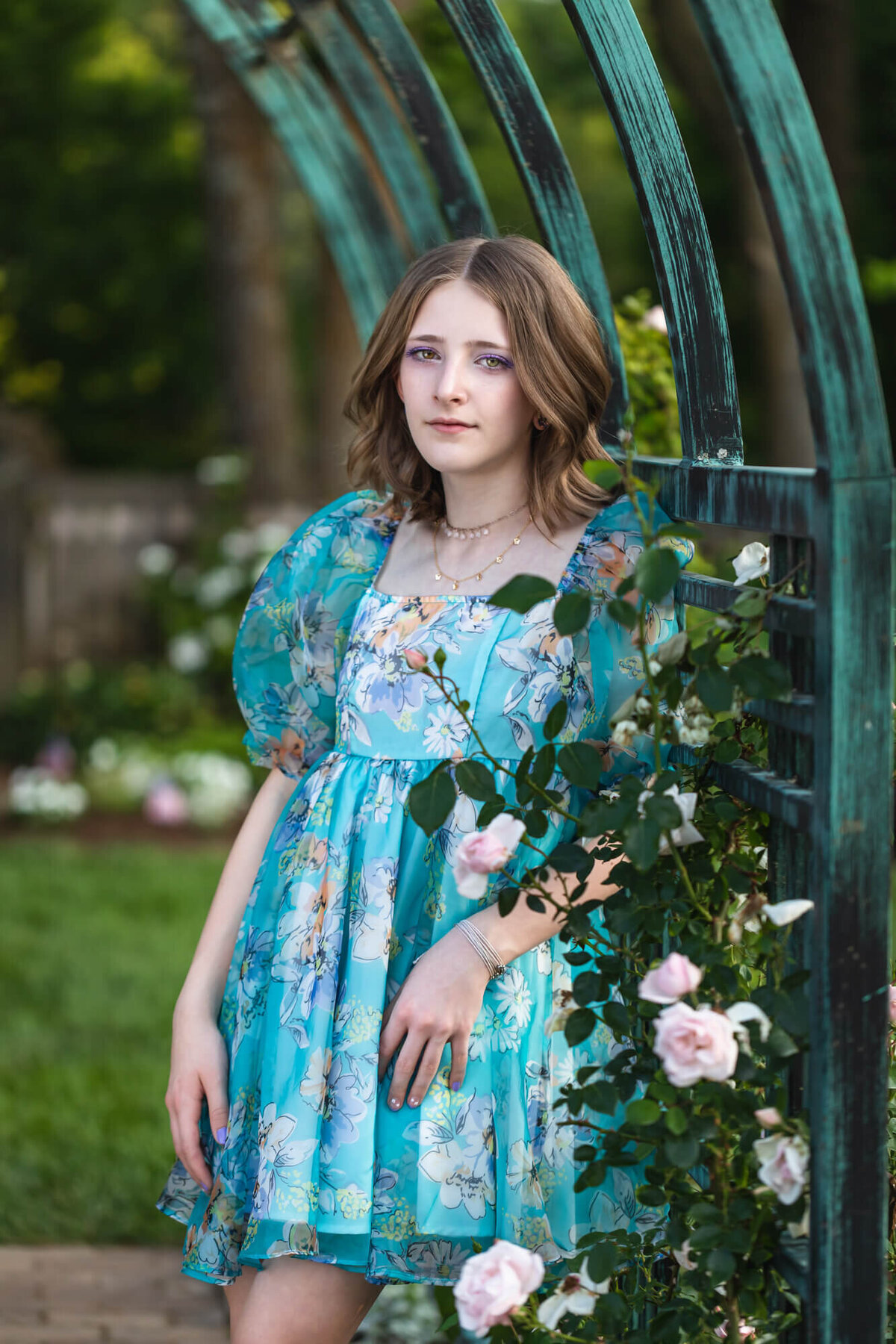 Lovely teenage girl in a blue puff sleeve dress leaning on a garden trellis surrounded by pale pink roses. Captured by Springfield, MO teen photographer Dynae Levingston.