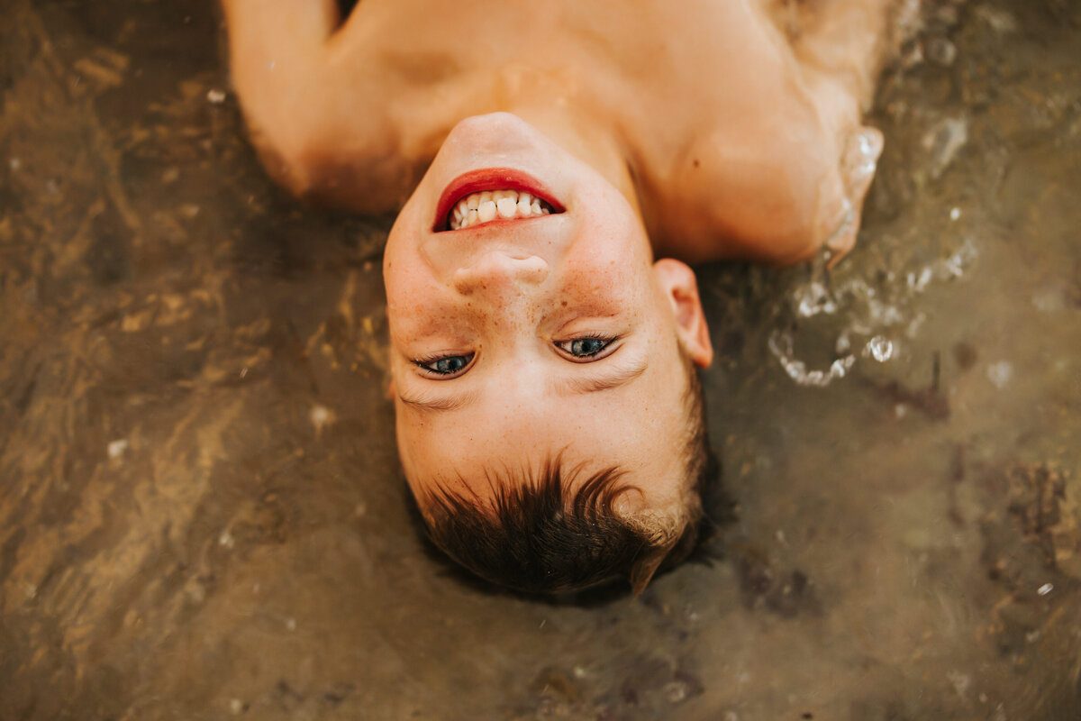 a boy laying in the ocean during a photo session splashing in the water