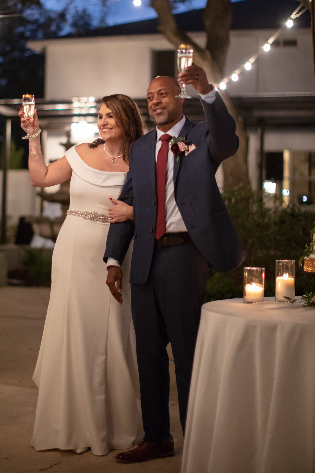 bride in scoop neck dress and groom with maroon tie toasts under string lights at San Antonio wedding Gardens at West Green