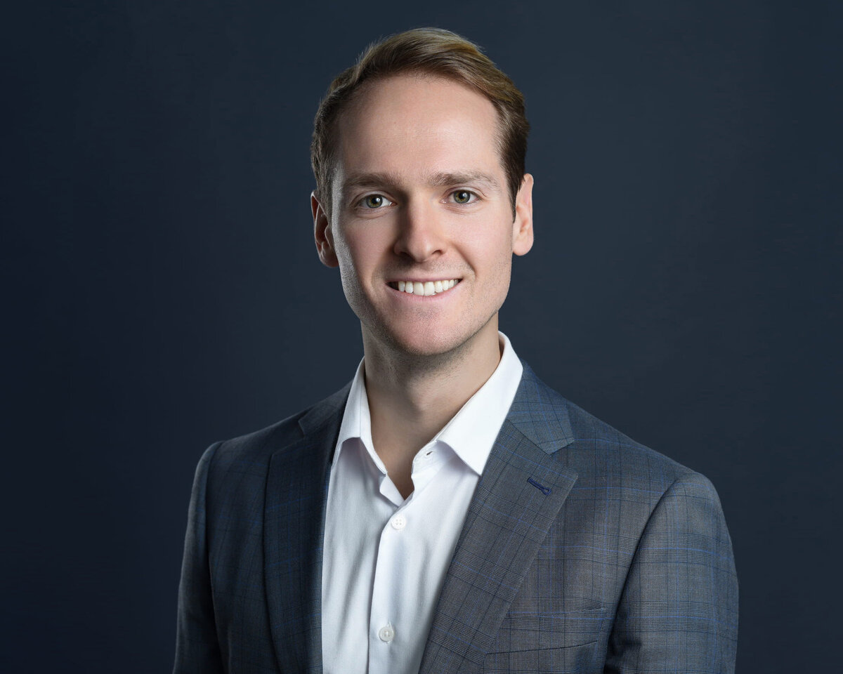 Professional headshot of an individual dressed in a dark grey checkered suit jacket and white dress shirt, posed against a rich dark blue backdrop. The subject’s polished look and confident expression make this an ideal branding photo for corporate and business professionals