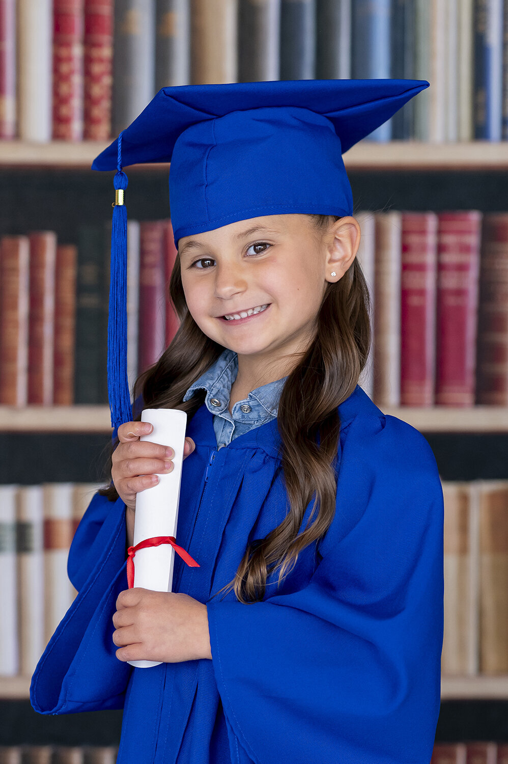 Preschool graduate holds a diploma in her hands while dressed in a blue cap and gown standing infront of a bookcase.