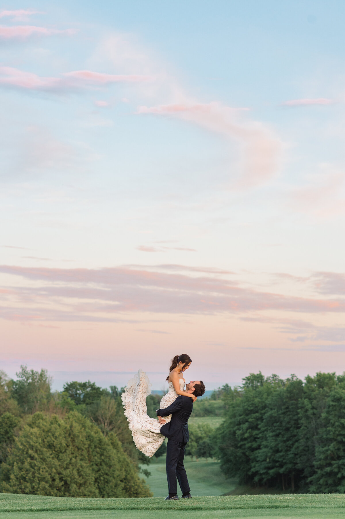 A wide colour photo of a bride in her grooms arms with her legs kicking behind her while he spins her at their backyard wedding at sunset