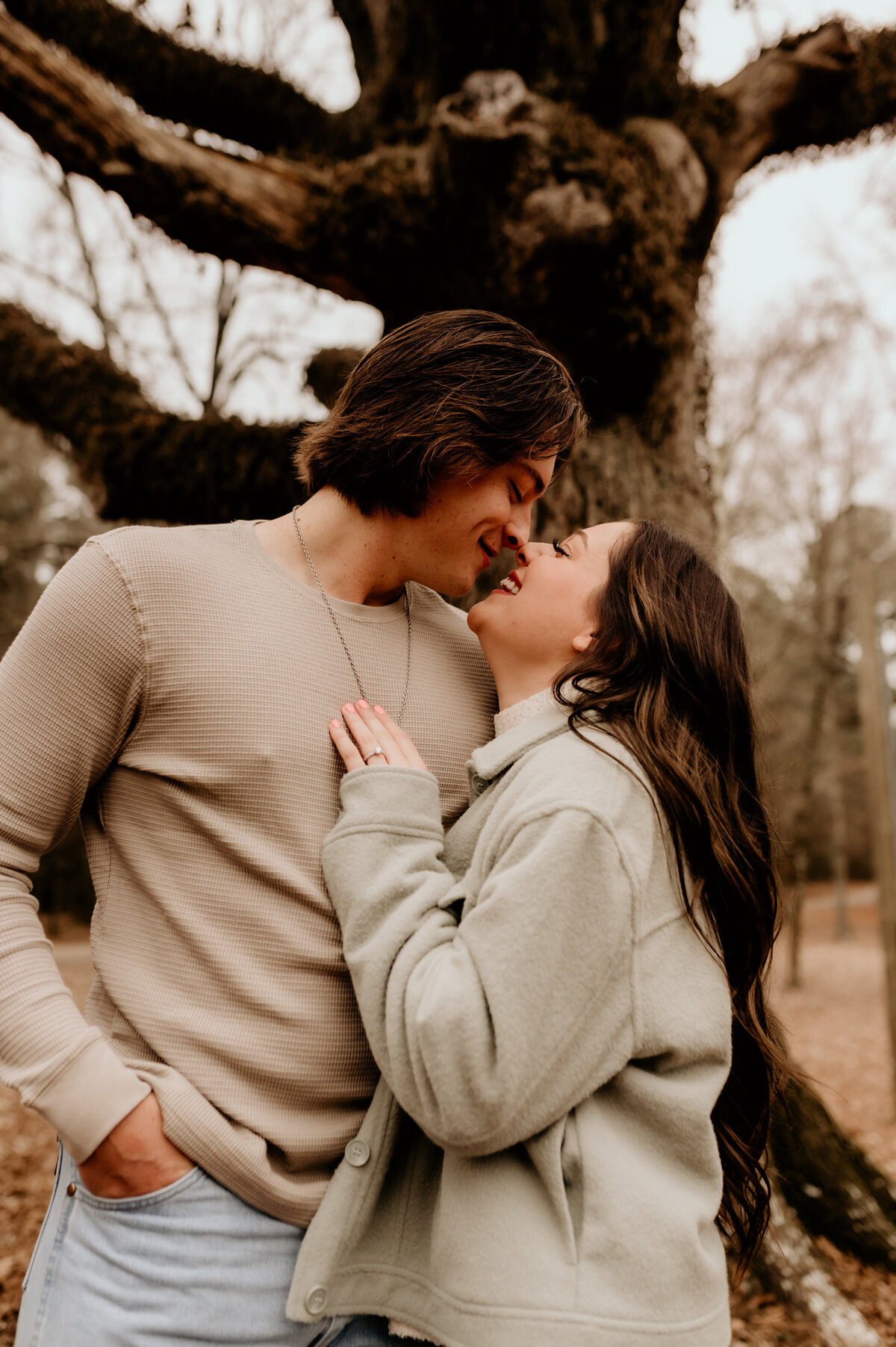 man leaning down to kiss his fiance as she laughs and smiles with a tree behind them