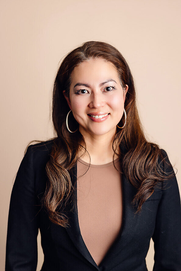 Woman with hair tucked behind ears smiles in front of tan background.