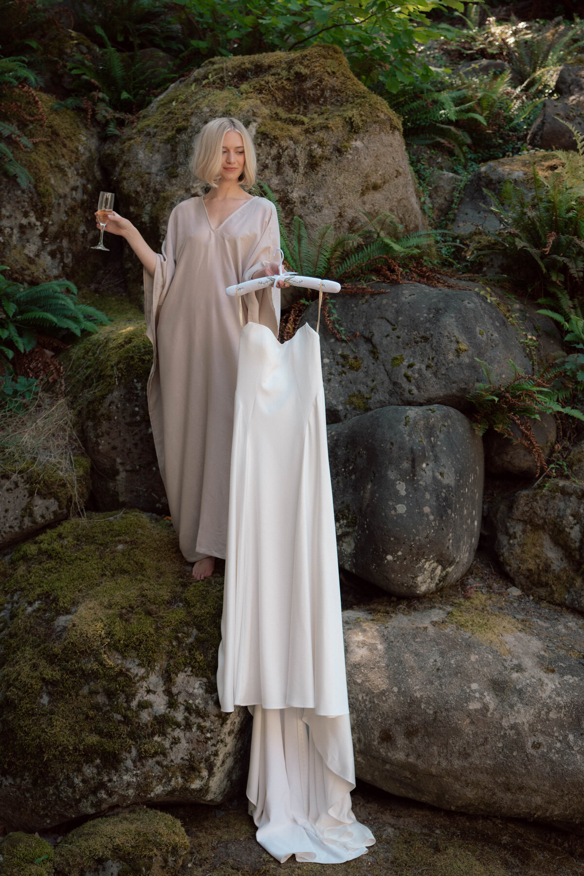 Bride in taupe robe holding wedding gown on hanger against mossy rocks