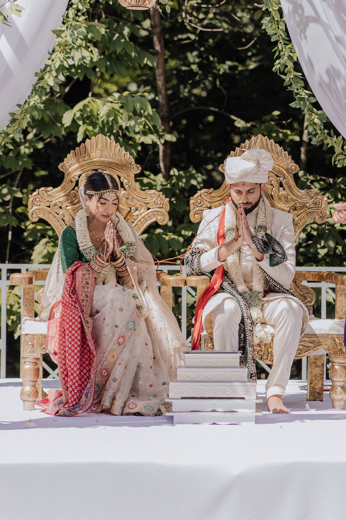 Hindu wedding ceremony photo of the bride and groom seated on ornate gold chairs, hands folded in prayer during their traditional rituals outdoors.