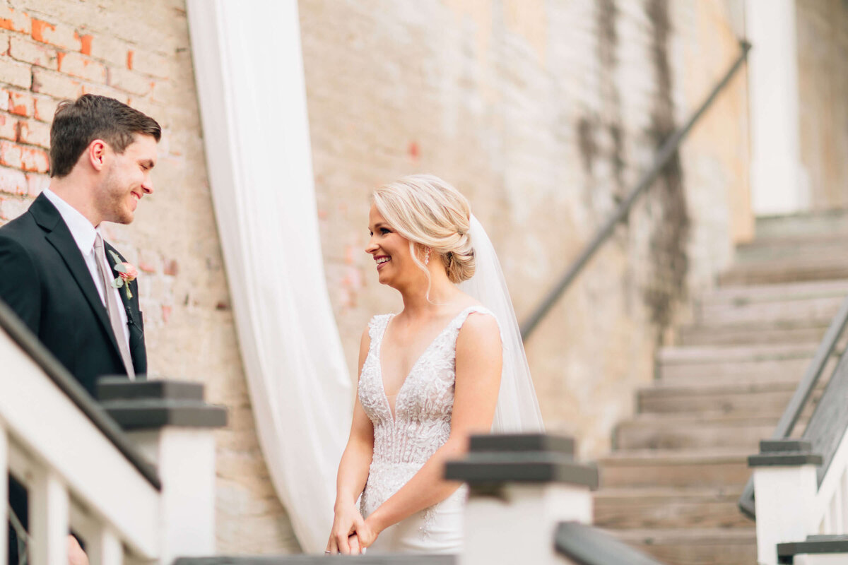 A bride in a white dress and veil smiles at a groom in a black suit and tie during their first look. They stand next to a brick wall and wooden stairs, sharing a joyful moment together.