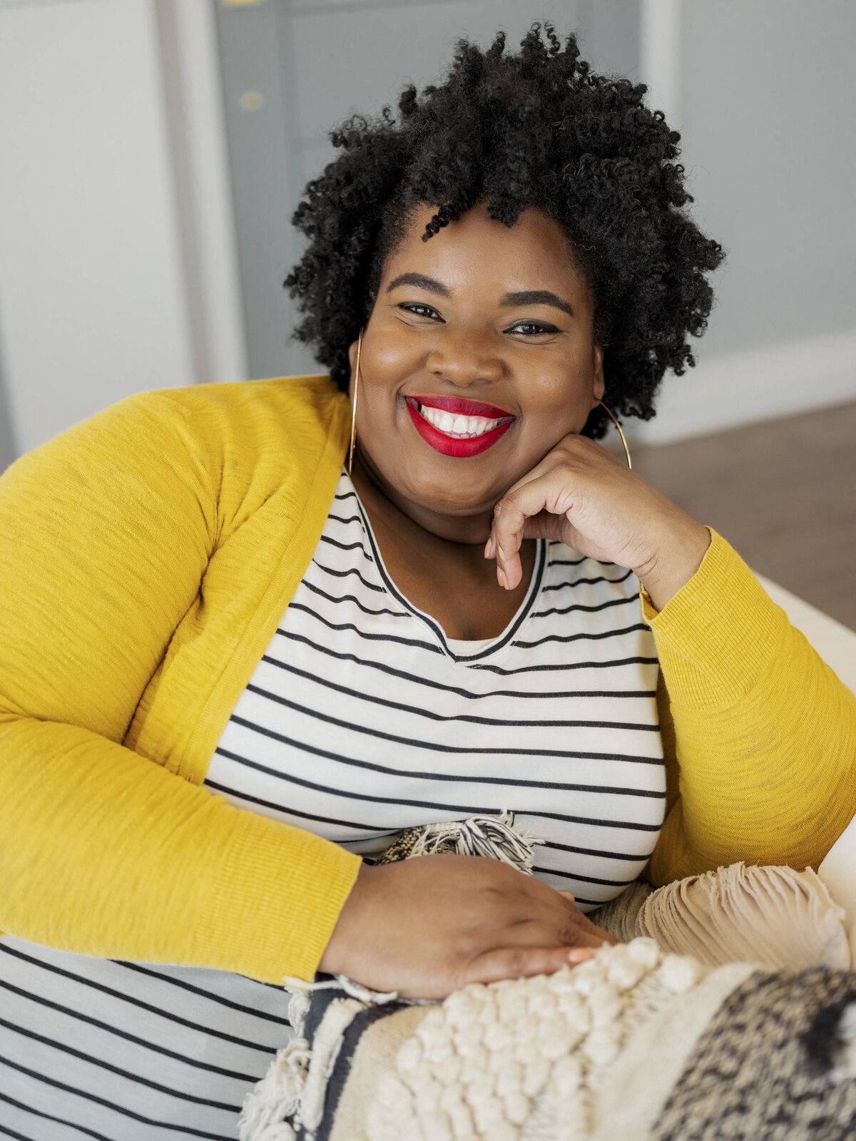 Black woman in striped shirt and yellow cardigan smiling while sitting on a couch during NYC brand shoot.
