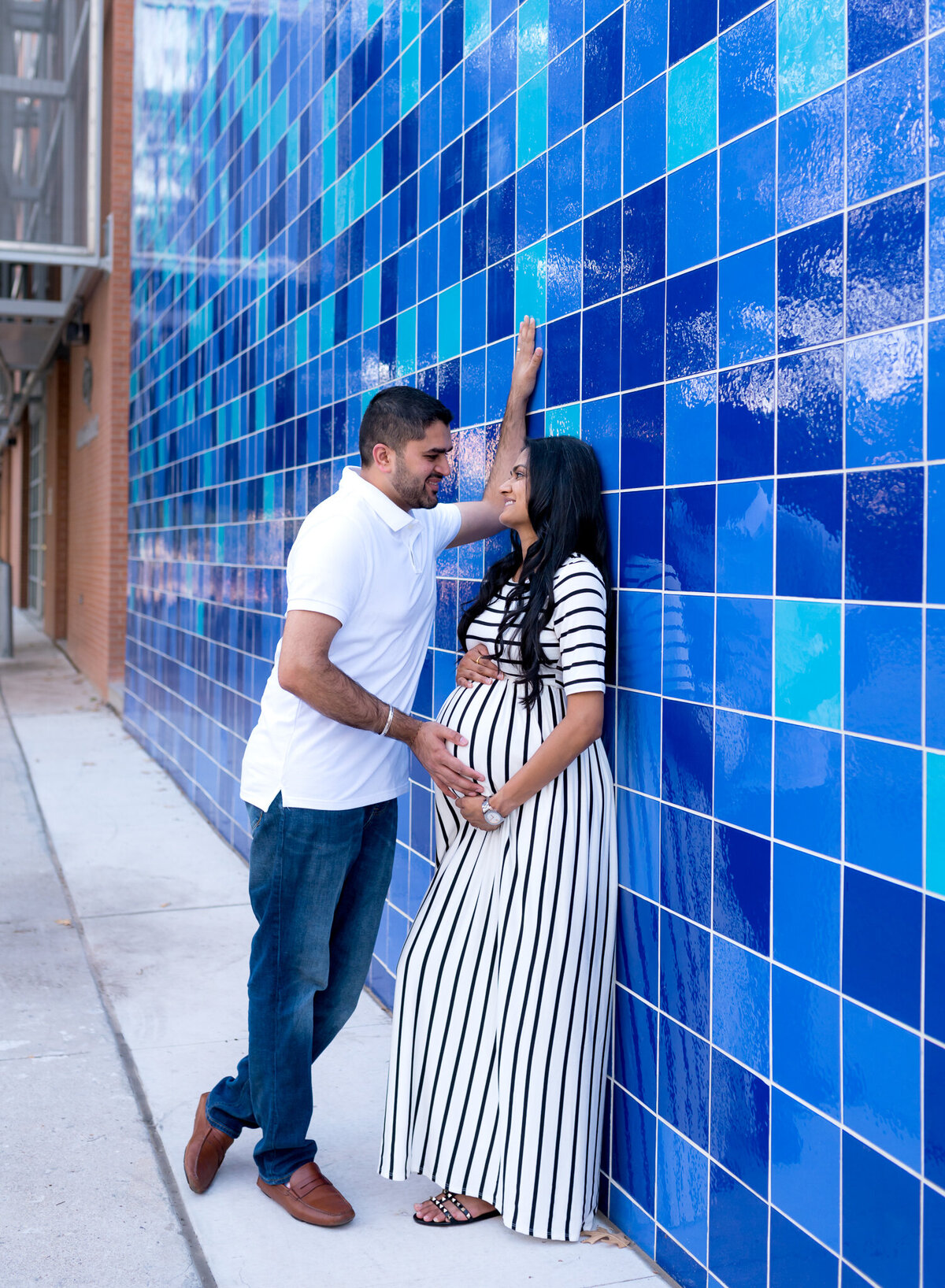 Urban maternity portrait in downtown Austin with a couple smiling and leaning against a bright blue tile wall. The mom-to-be wears a black and white striped dress.