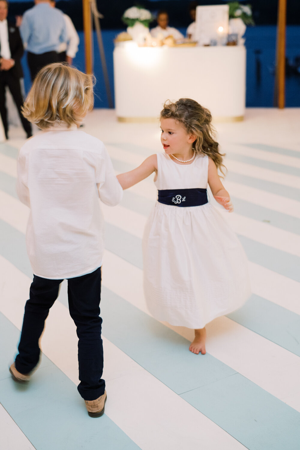 The photo was taken at The Hamptons, New York, waterfront estate tented wedding by photographer Bonnie Sen, with videographers East West Films capturing the moment. In the photo, two young children are dancing together at the formal event. One is a boy with blond hair, wearing a white shirt and black pants, while the other is a girl with curly hair in a white dress with a navy blue sash. A bar can be seen in the background.