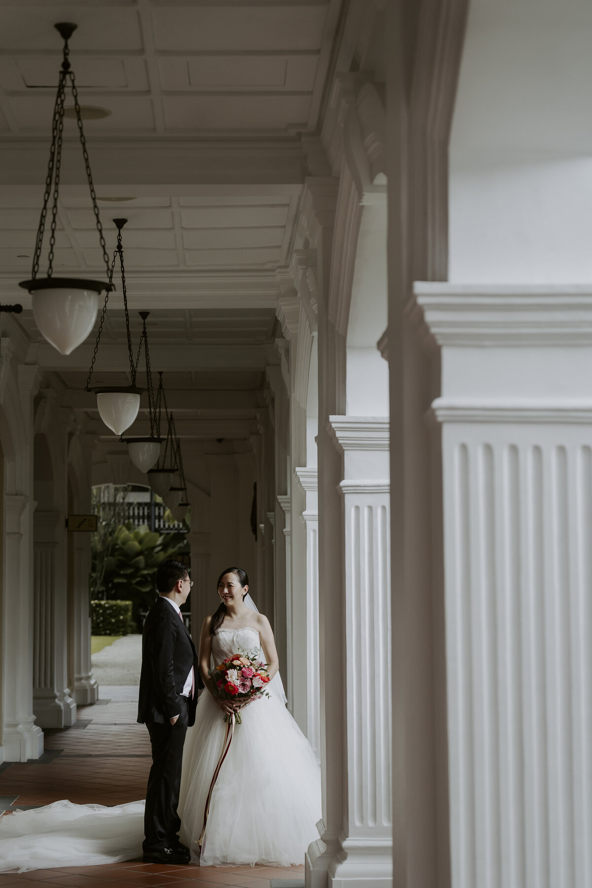the bride and groom smile at each other between white pillars at the raffles hotel