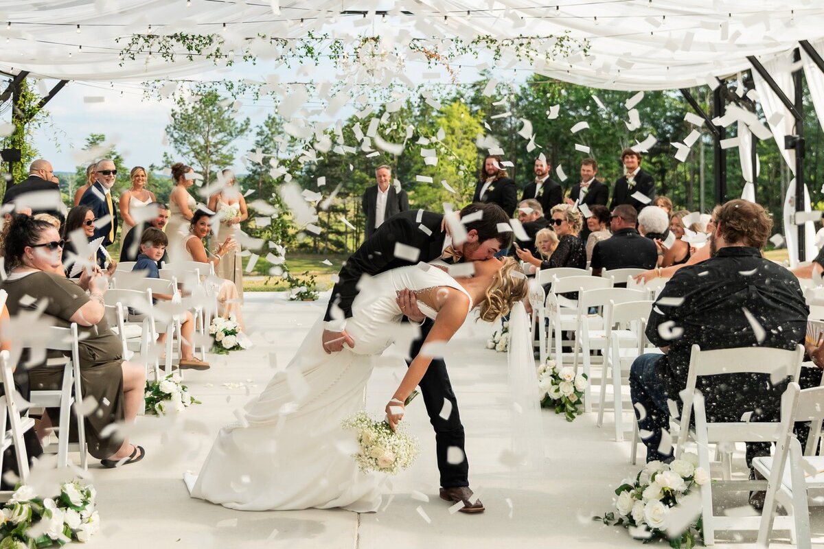 A groom dips his bride into a dramatic kiss under a white canopy during their ceremony, just as white confetti showers down around them. The bride clutches her bouquet of baby’s breath, and their guests cheer from either side.