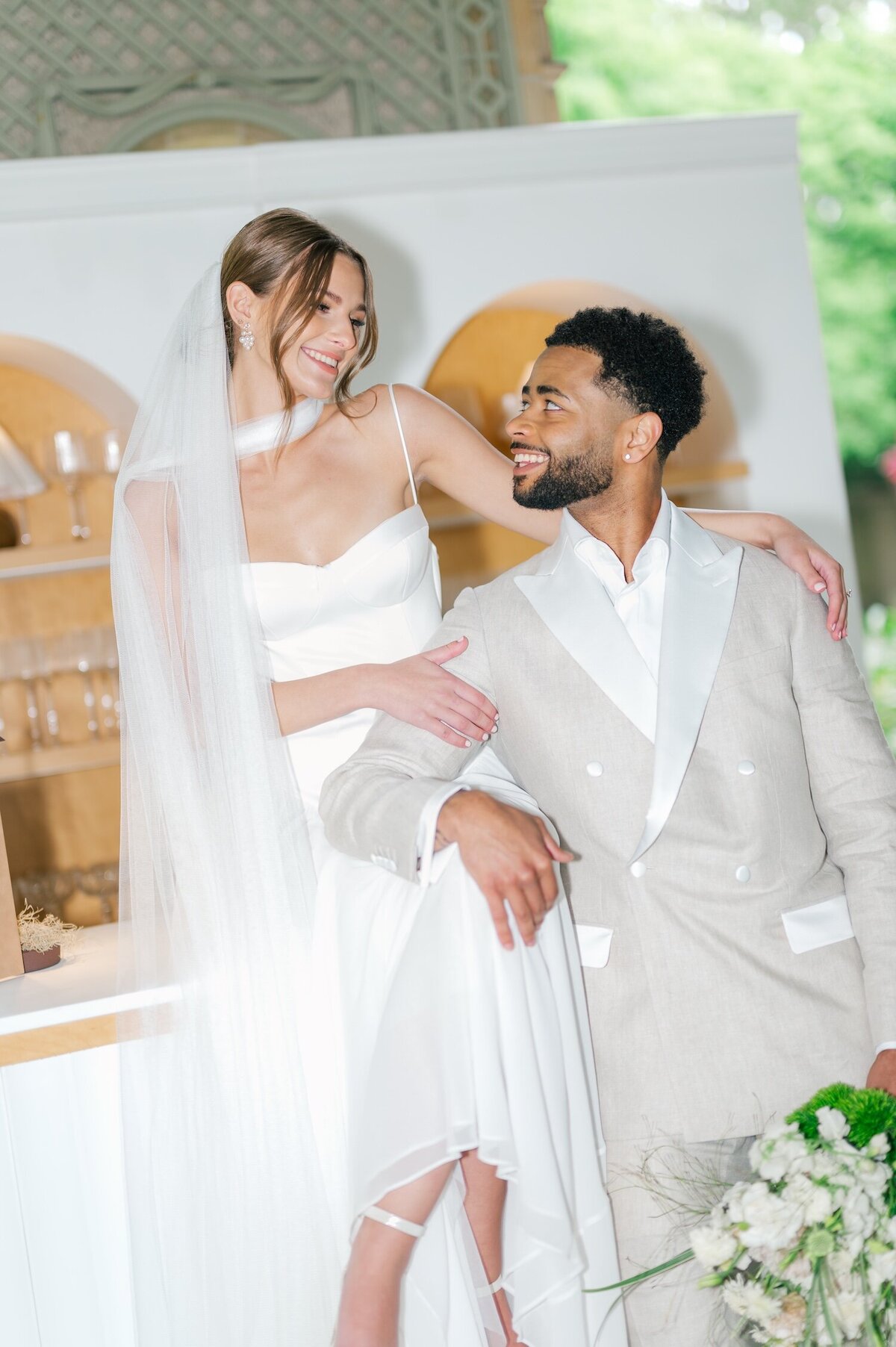 A bride sits on top of a bar and her groom leans against her and laughs. 