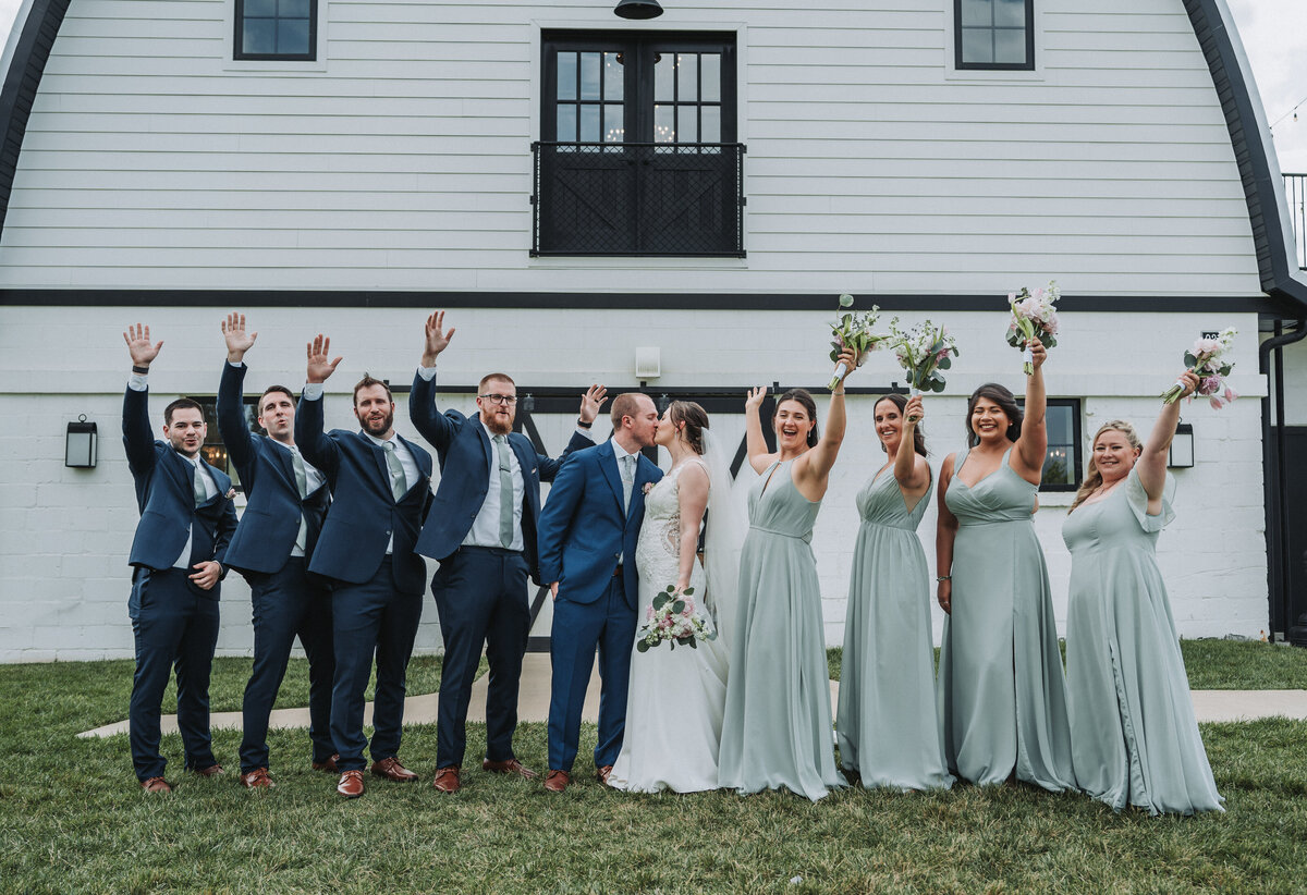 Bride and groom kiss while their wedding party cheers in front of the white barn at Sweeney Barn in Manassas, Virginia.