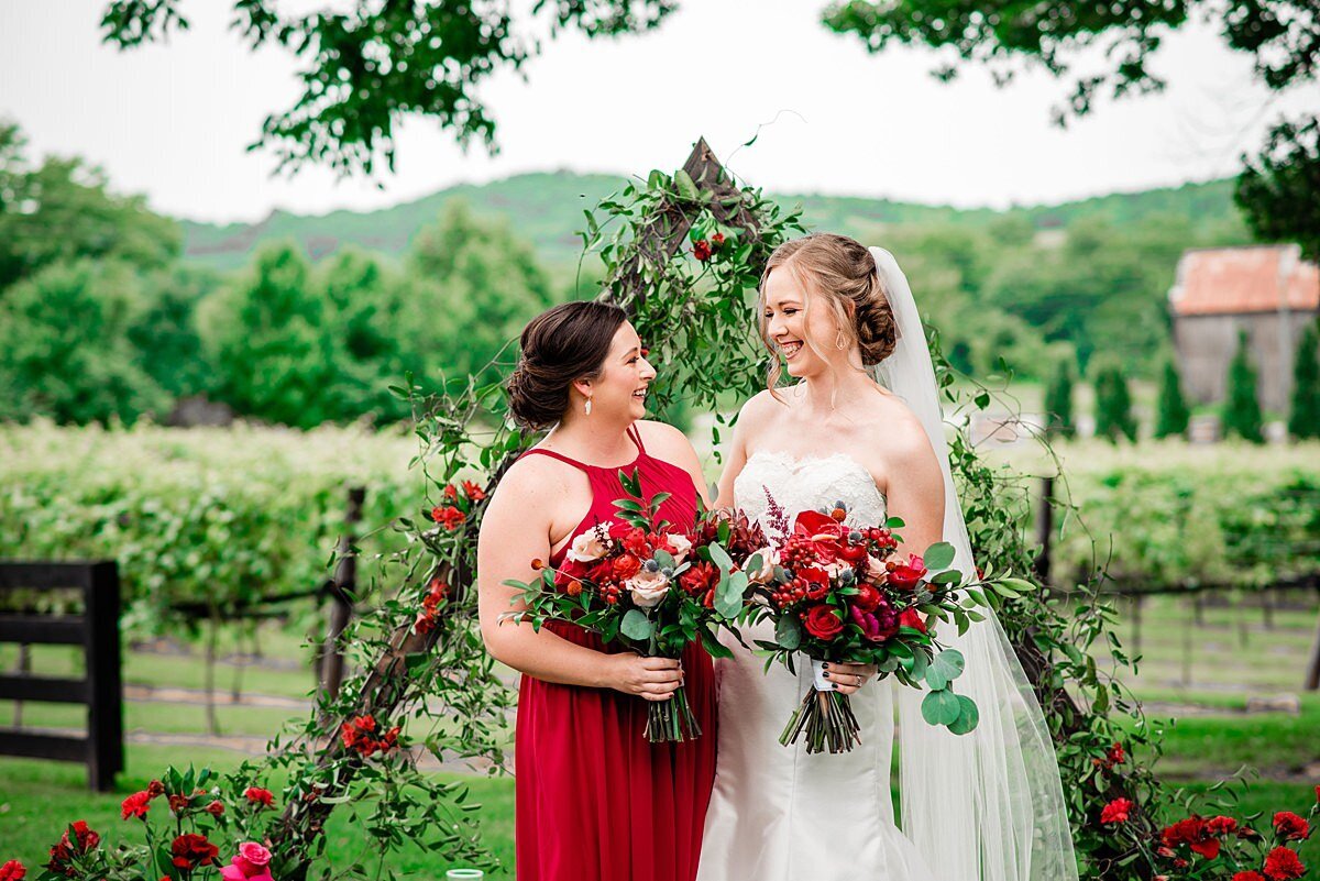 The bride, in a silk strapless wedding gown with a lace top featuring a sweetheart neckline laughs with her maid of honor dressed in a red chiffon halter top bridesmaid dress. The bride and maid of honor are holding bouquets of red roses, blush roses, red anthurium, red anemones, protea, blue thistle, hypericum berries, dianthus and assorted greenery as they stand in front of a triangular wedding ceremony arbor covered in smilax vine, red roses, and red carnations with the wine vines of Arrington Vineyards in the background.