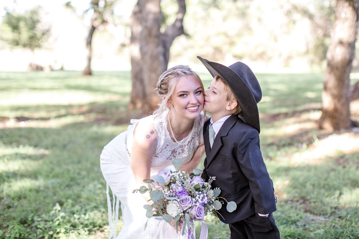 ring bearer with cowboy hat kisses bride on cheek as she bends down holding lavender bouquet