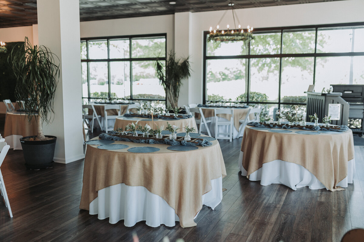 Elegant reception setup at the Hampton Roads Event Center, featuring round tables with beige tablecloths and rustic centerpieces, surrounded by large windows with views of greenery. Shot by Quinn Photography LLC.