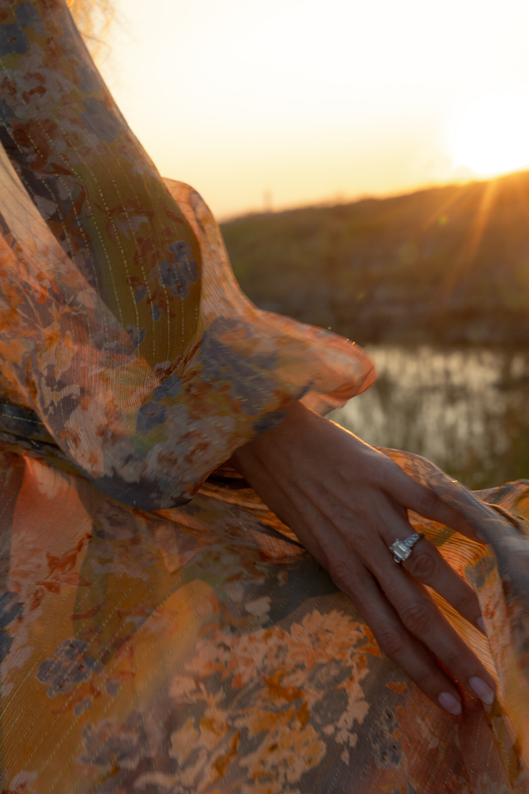 Woman's hand with engagement ring is holding on to orange dress with flower patterns. Sun is setting in the background and provides warm color. Engagement photo taken in Charleston.