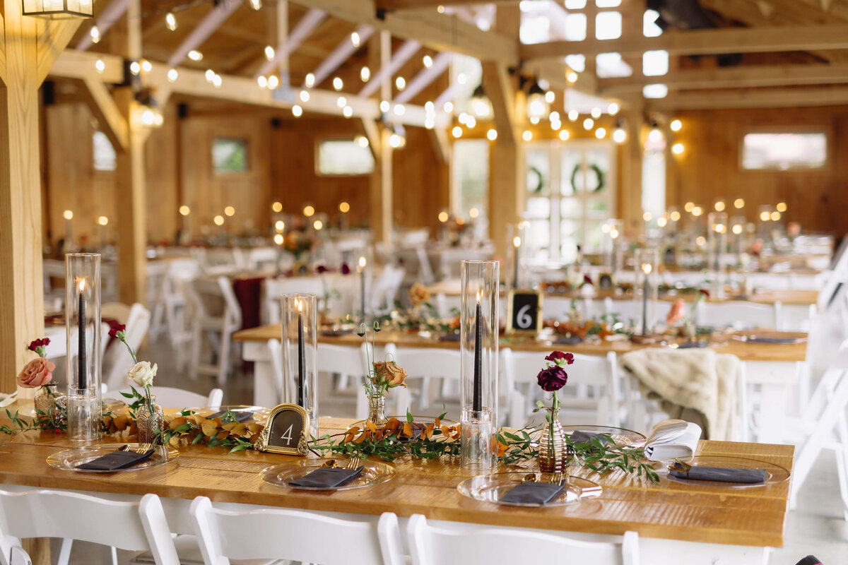 Charlottesville wedding photographer photographs reception hall at Layz S Ranch with string lights over head with wooden tables and white chairs with the tables decorated with glass, candles and greenery