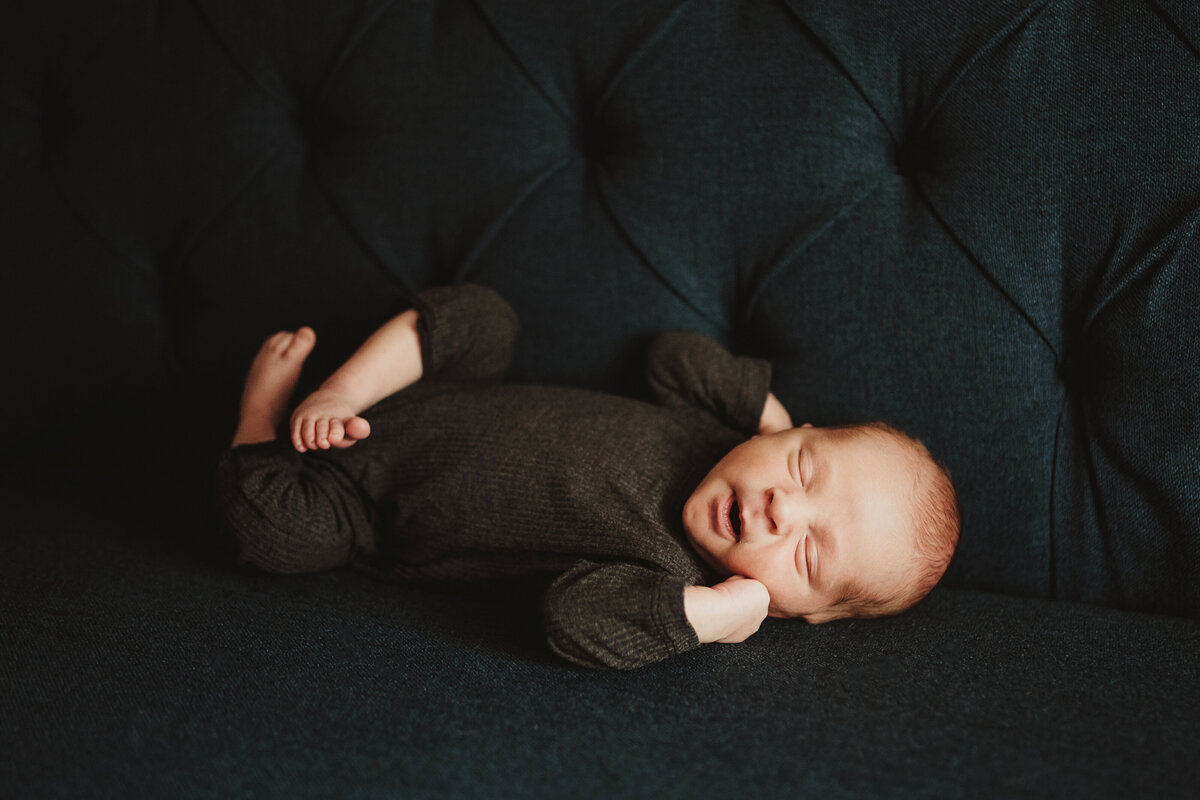 Baby boy in a dark gray romper peacefully sleeping on a navy blue couch.