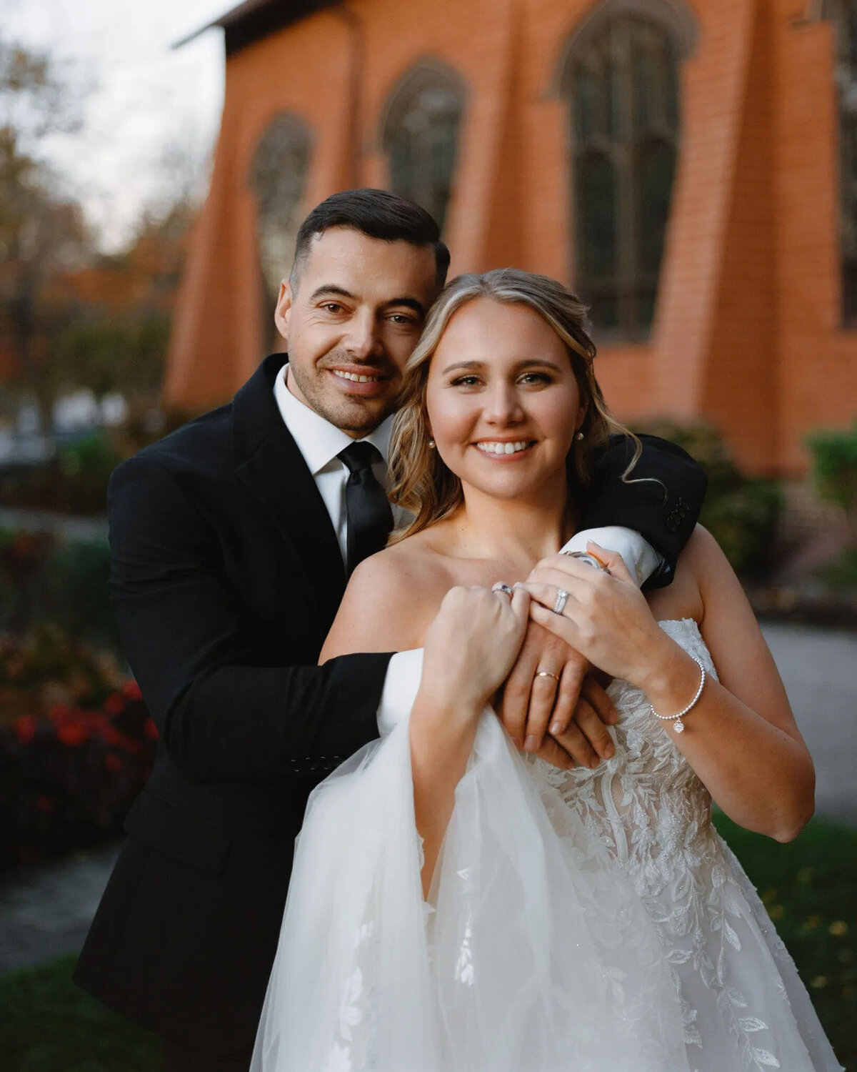A smiling bride and groom pose outside in front of a brown building with arched windows. Captured by a talented NJ wedding photographer, the groom hugs the bride from behind as they show off their rings, radiating joy in their formal wedding attire.