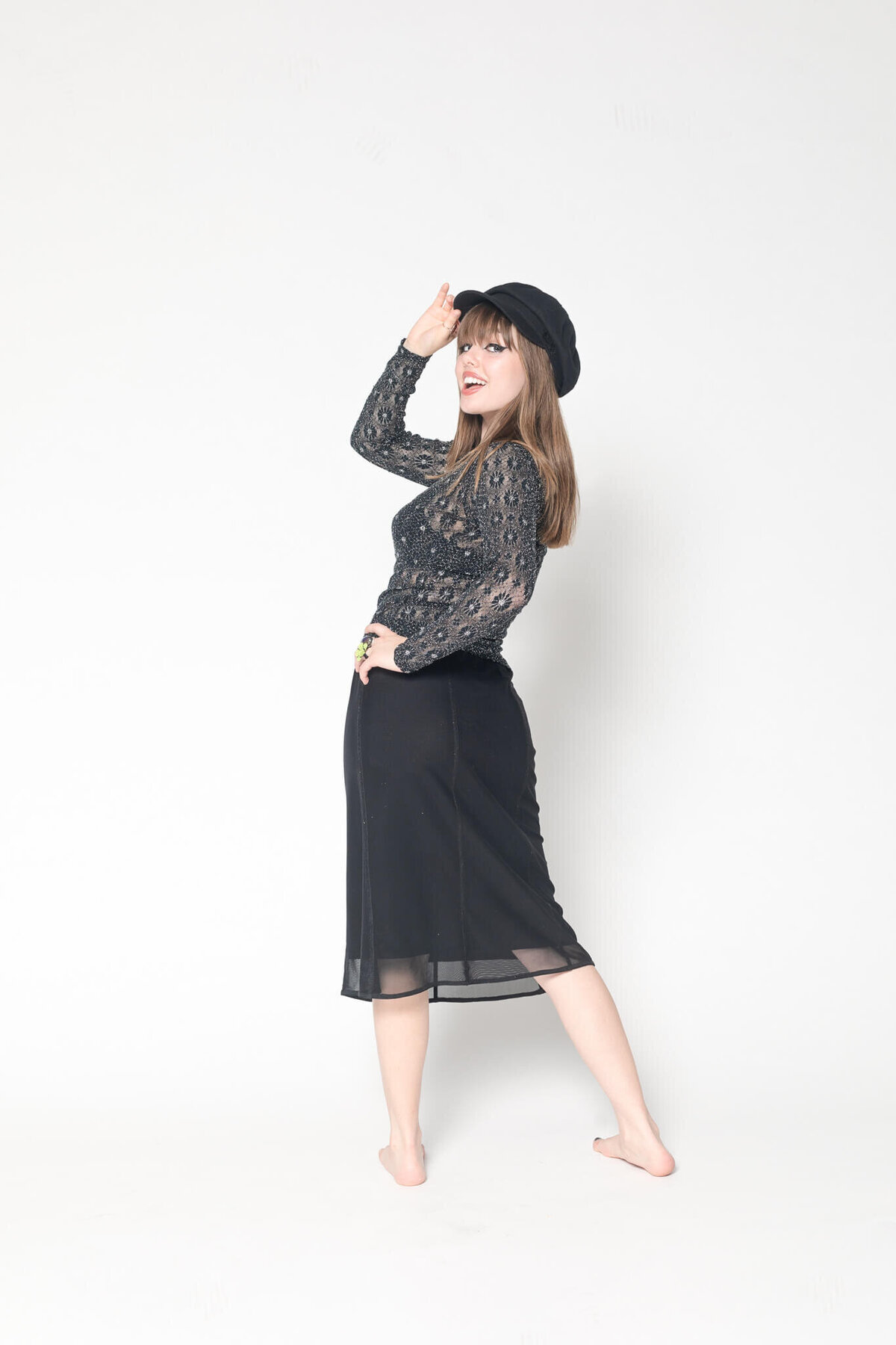 A person wearing a black lace top, black skirt, and hat poses against a white background. They are standing barefoot and have one hand touching the brim of the hat, smiling while looking to the side.