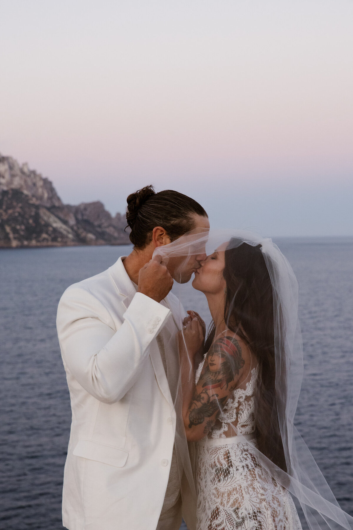 Husband and wife kissing each other under a veil in front of Es Vedra on Ibiza during an elopement photoshoot