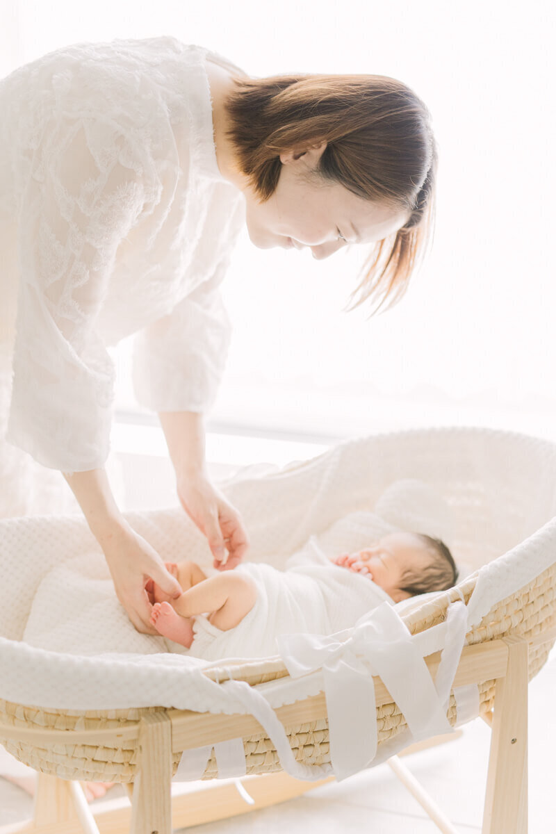 オシャレなゆりかごの中で寝ている赤ちゃんの足を触りながら微笑んでいるママが美しい、ナチュラル・ニューボーンフォト