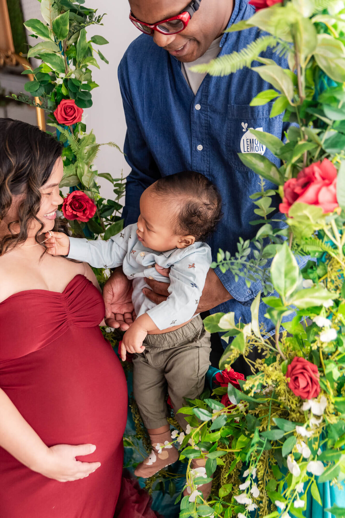 A pregnant woman and her husband joyfully cradle their baby, surrounded by a vibrant floral display in Youngstown Ohio photography session