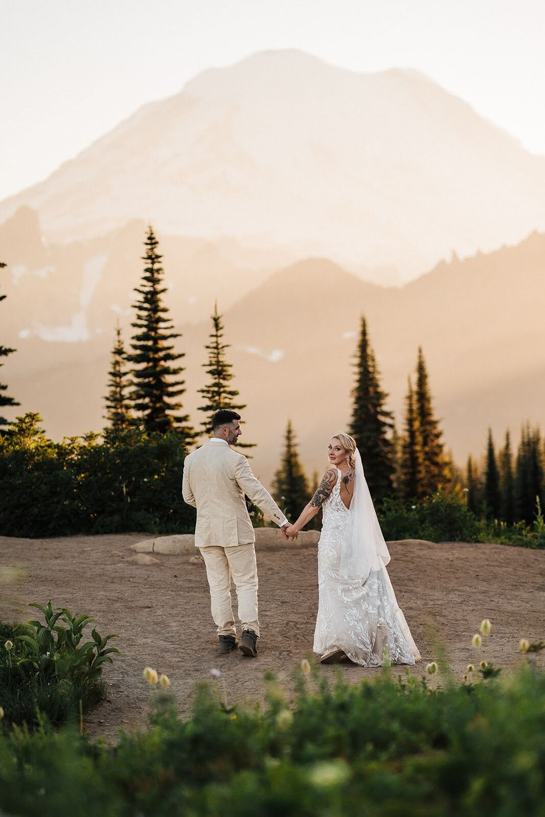 Bride & groom holding hands and looking back as they walk toward Mount Rainier at sunset on the Naches Peak Loop trail!