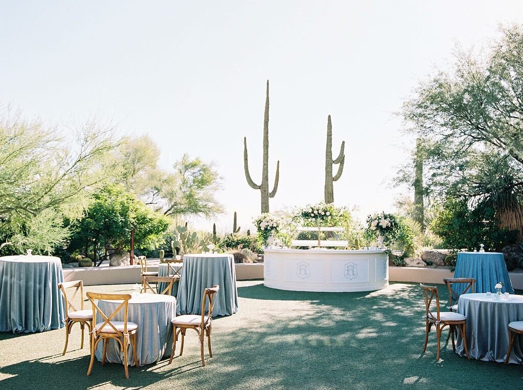 A beautifully designed outdoor wedding bar setup with elegant floral arrangements and soft blue linens, set against the stunning desert landscape.