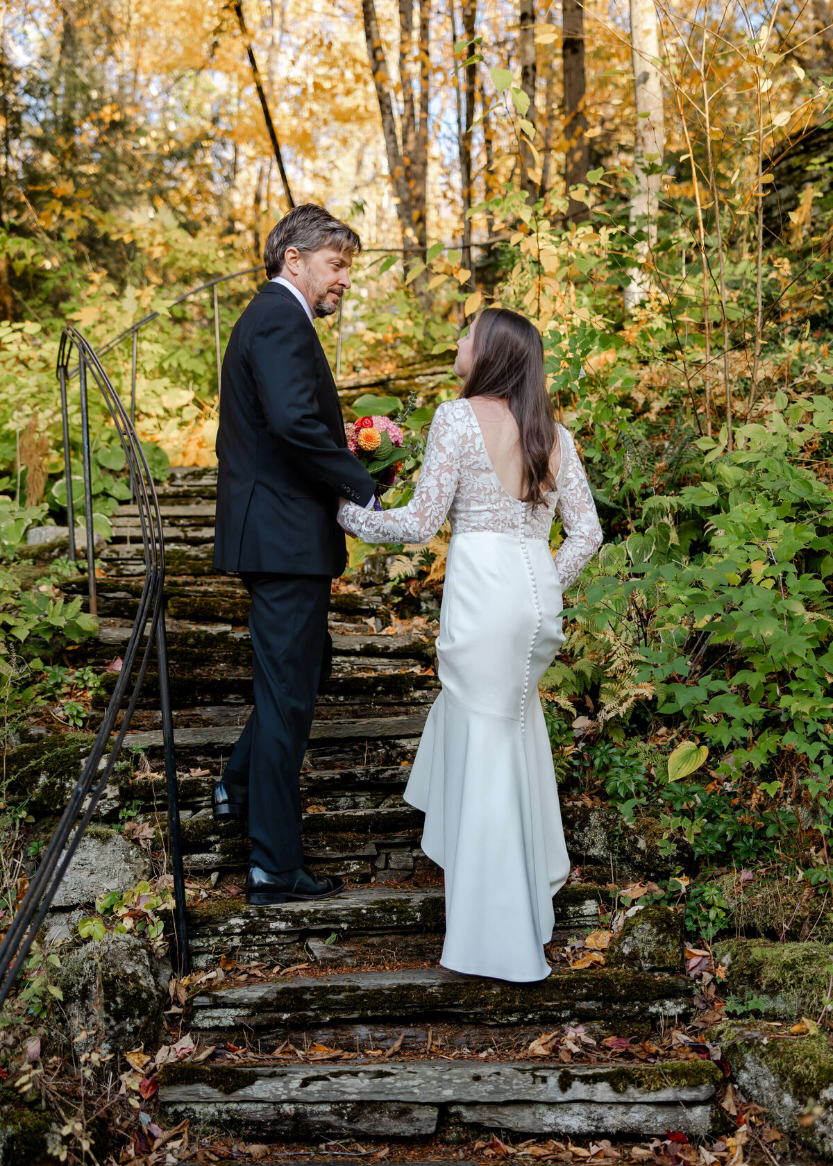 A groom gently helps his bride ascend a rustic wooden stairway leading into a serene Vermont forest, capturing a tender moment during their intimate wedding ceremony