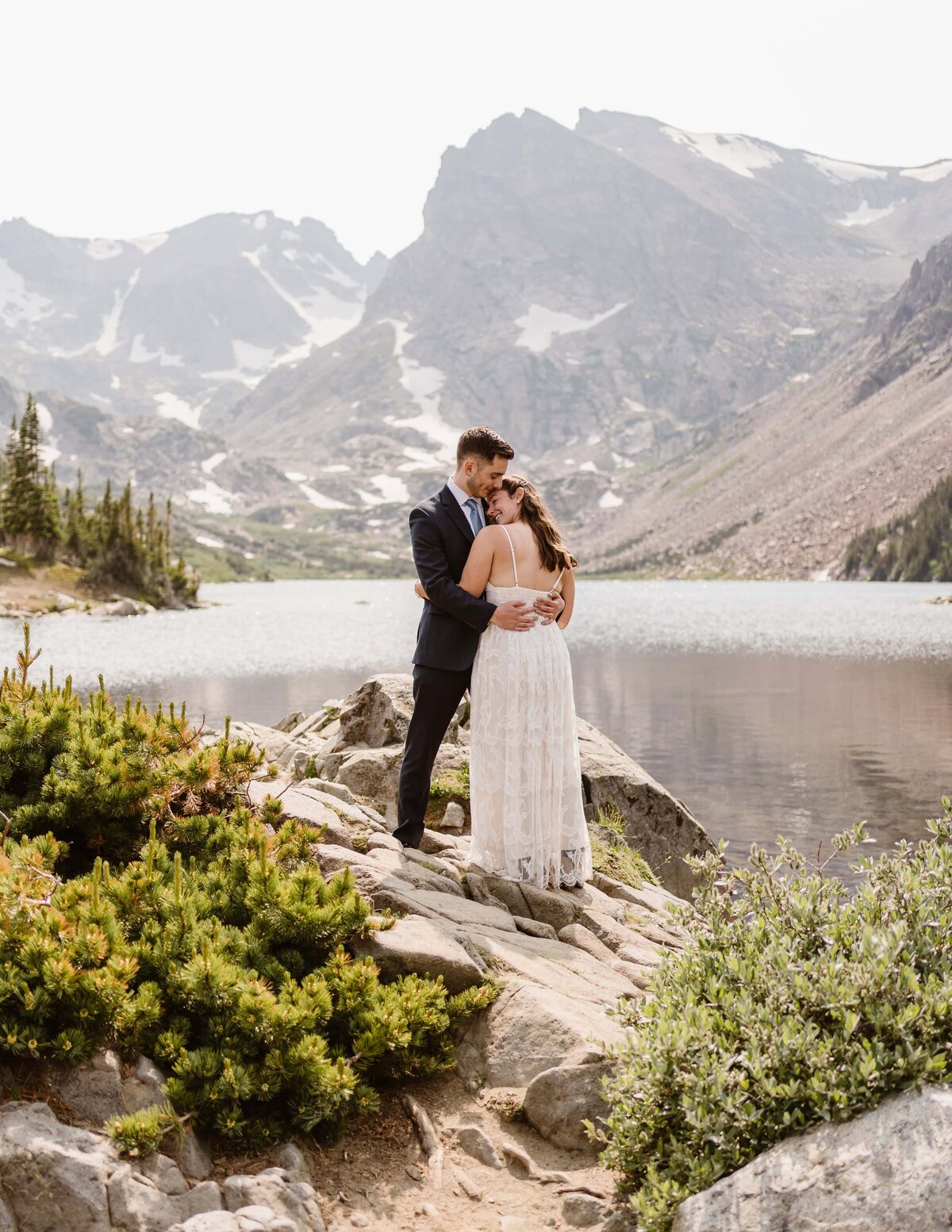Bride and groom embrace on rocky lakeshore with towering snow-dusted peaks in the background during their Lake Isabelle elopement.