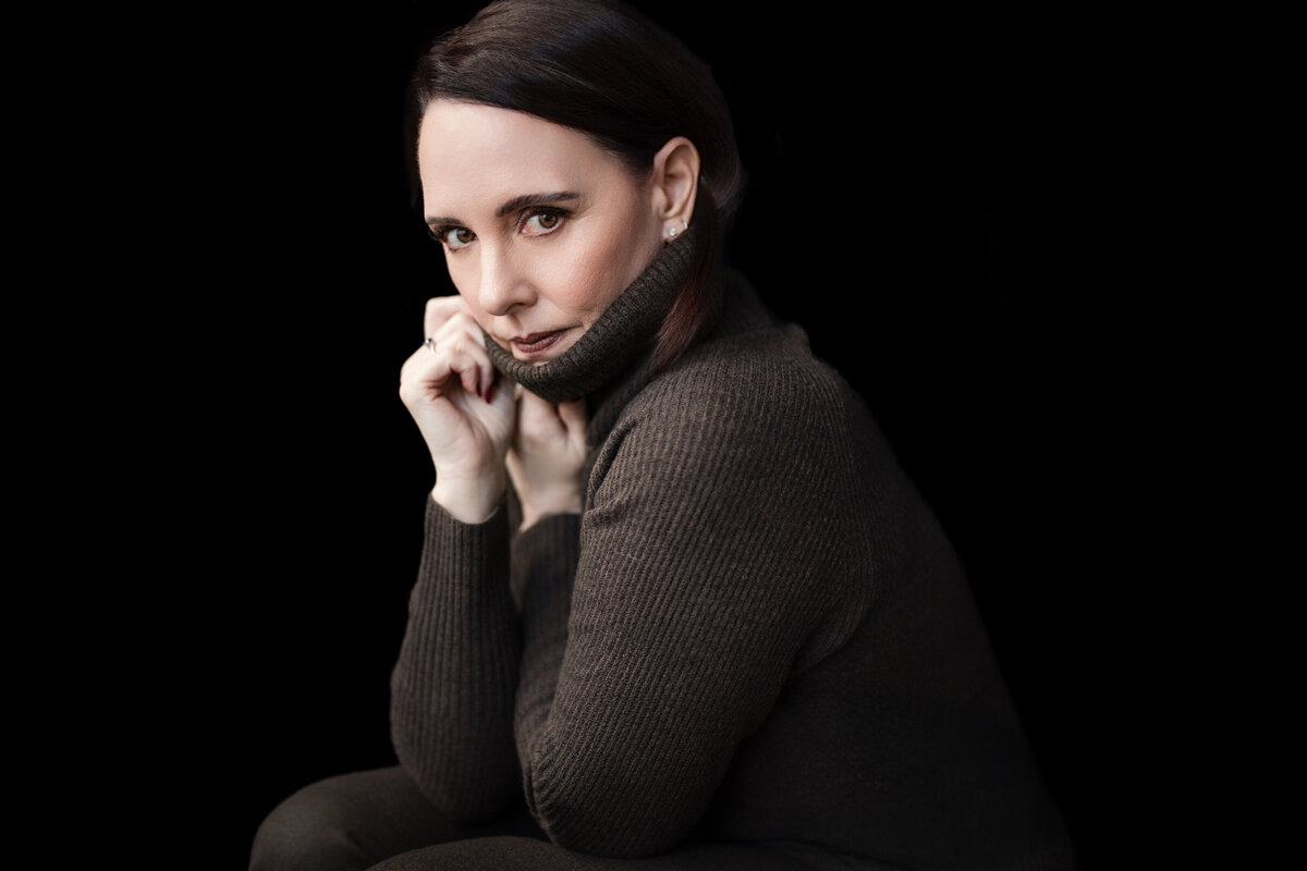 Person seated against a solid black background, wearing a dark ribbed turtleneck sweater pulled up to their chin. They look directly at the camera with a calm, intense expression and softly lit face, creating a dramatic and intimate headshot.
